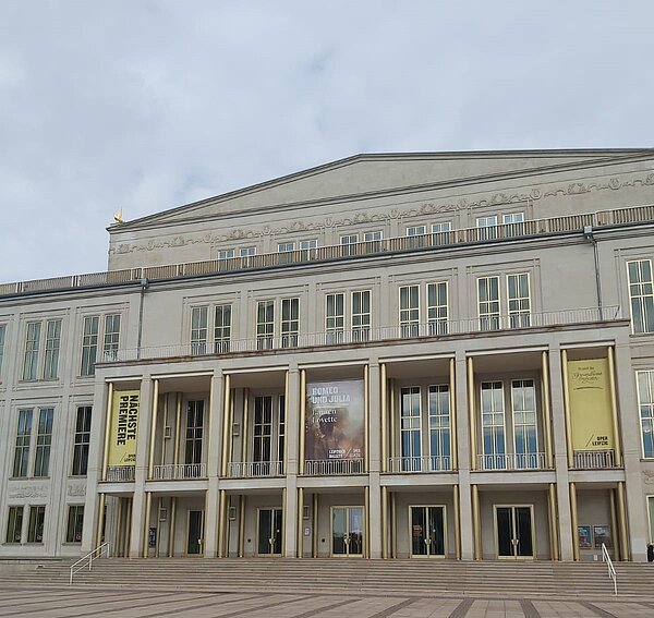 Ein klassisch-modern gestaltetes Gebäude. Die Fassade ist symmetrisch und in einem hellen Ton gehalten, das Gebäude hat großen Fenster und Säulen im Eingangsbereich. Auf Bannern vor einigen Fenstern steht unter anderem „Oper Leipzig“.