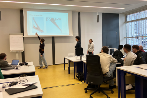 Foto des Seminarraums mit kleiner Gruppe Studierender, die am Beamer-Bild über konstruiertes Bauteil für Regalhalterung reden