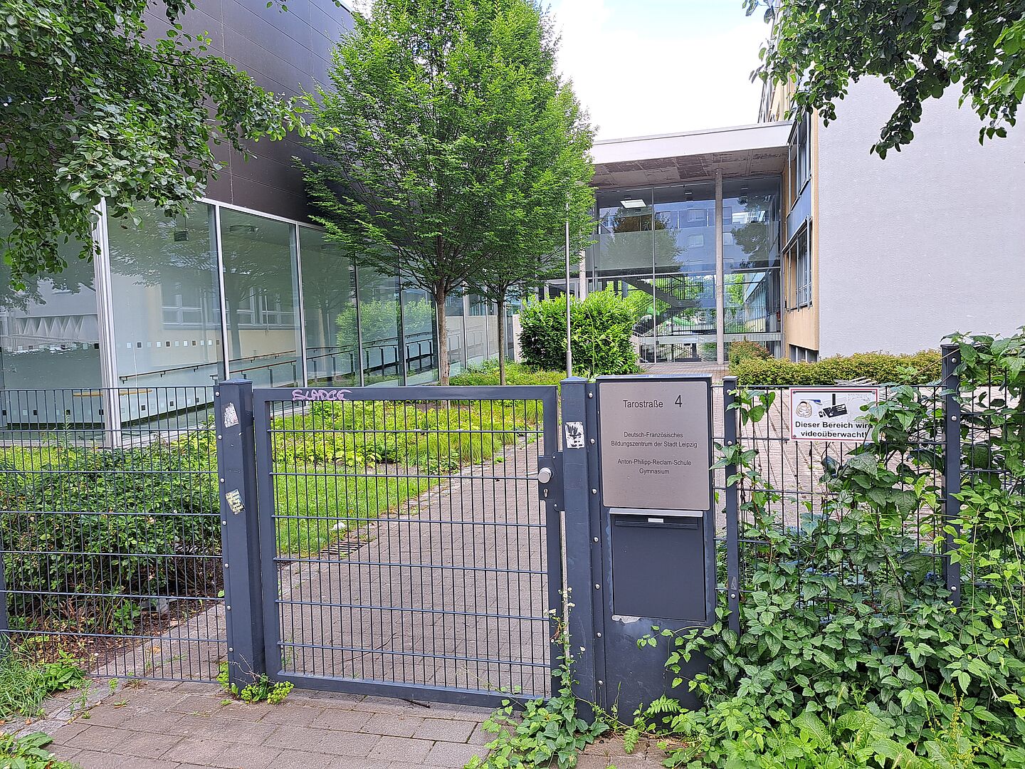 Das Bild zeigt den Eingangsbereich der Anton-Philipp-Reclam-Schule, ein modernes Schulgebäude mit großen Glasfassaden und abwechselnd dunklen und hellen Bauelementen. An einem Metalltor im Vordergrund steht die Adresse "Tarotstraße 4". Das Schulgelände ist von grünen Bäumen und Büschen umgeben, und ein Schild informiert über die Videoüberwachung des Bereichs.