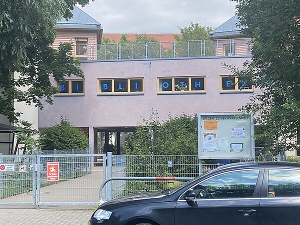 Leipziger Stadtteilbibliothek Wiederitzsch Ein grau-rosanes Gebäude mit einigen kleinen Fenstern. In den Fenstern steht jeweils ein Buchstabe des Wortes „Bibliothek“. Vor dem Gebäude ist ein silberner Metallzaun.