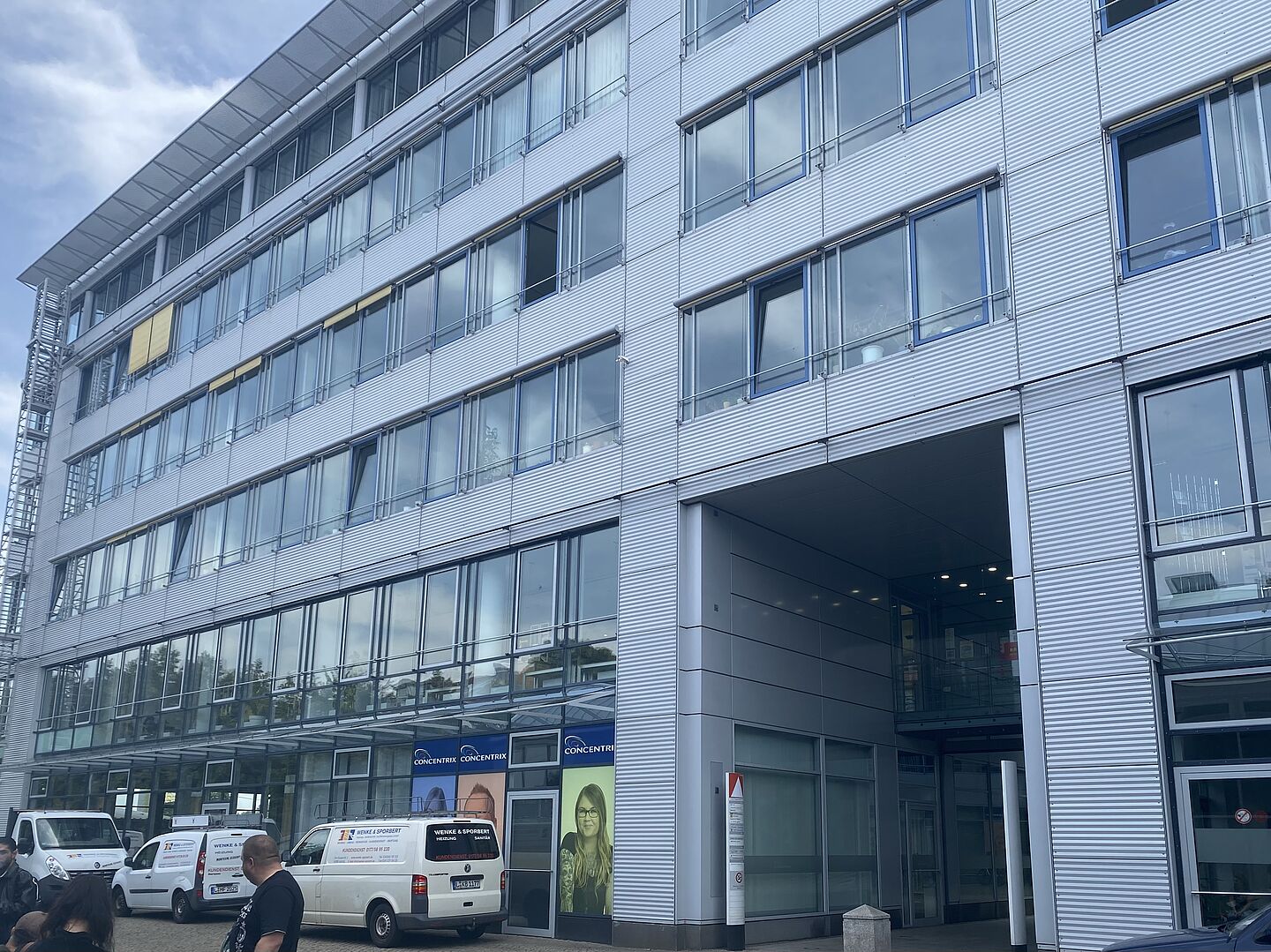 Das Bild zeigt ein modernes, mehrstöckiges Bürogebäude mit einer metallischen Fassade und zahlreichen großen Fenstern. Es hat mindestens fünf Stockwerke, und im Erdgeschoss gibt es einen überdachten Eingangsbereich. 