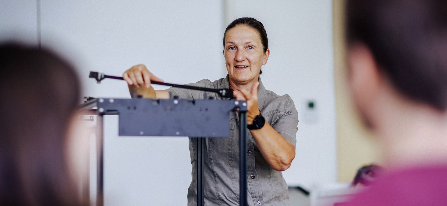 Eine Frau steht hinter einem schwarzen Gestell eines 3D-Druckers und erklärt Studierenden den Aufbau.