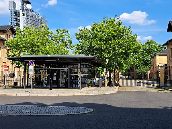 Ein kleines Glasgebäude, auf dem zweimal die Aufschrift „mdr“ steht. Daneben ist ein großes Metalltor, dass eine Zufahrt verschließt. Zu den Rändern sind sandfarben verklinkerte Häuser.
