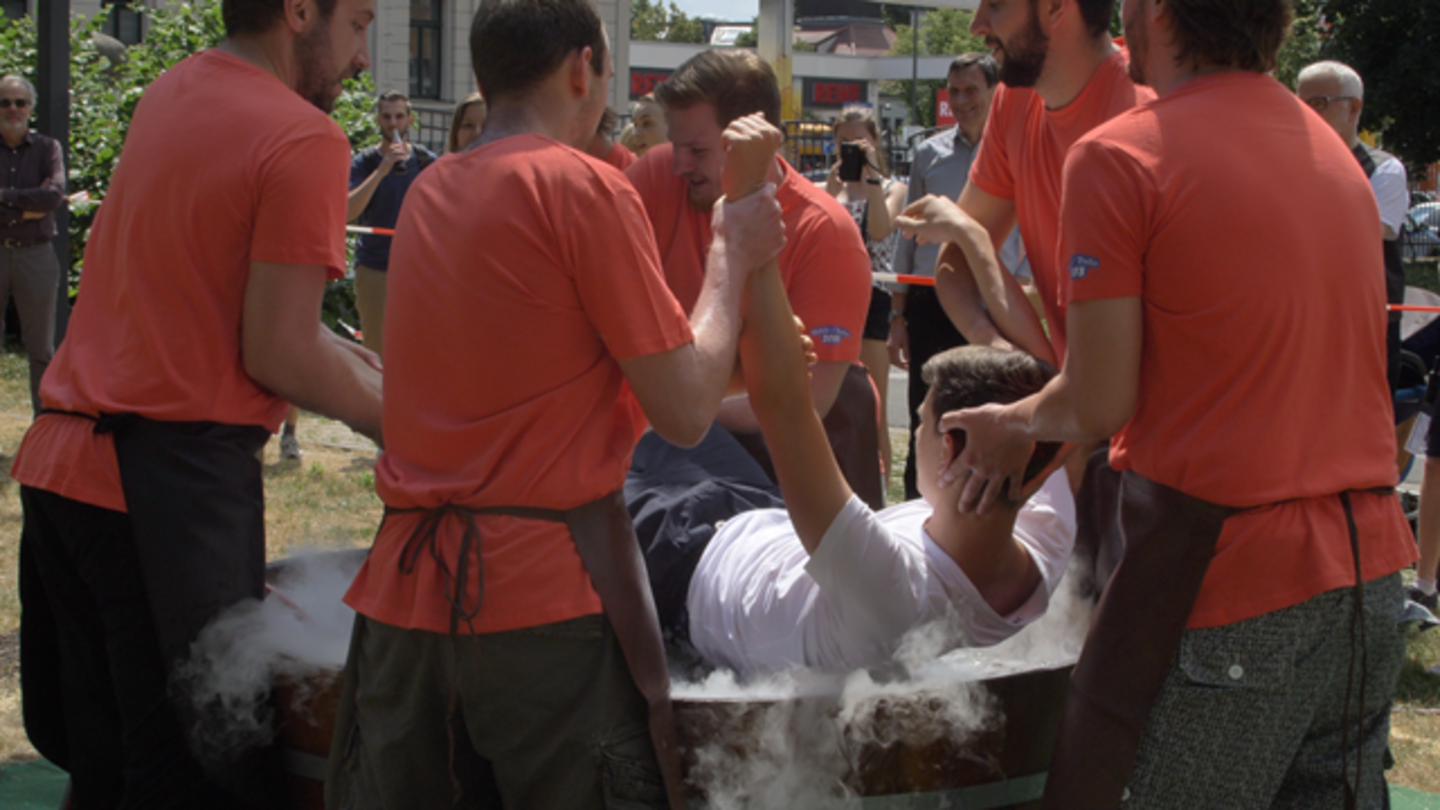 Mehrere junge Männer in roten T-Shirts legen einen jungen Mann in eine Badewanne. Das ist ein altes Aufnahmeritual.