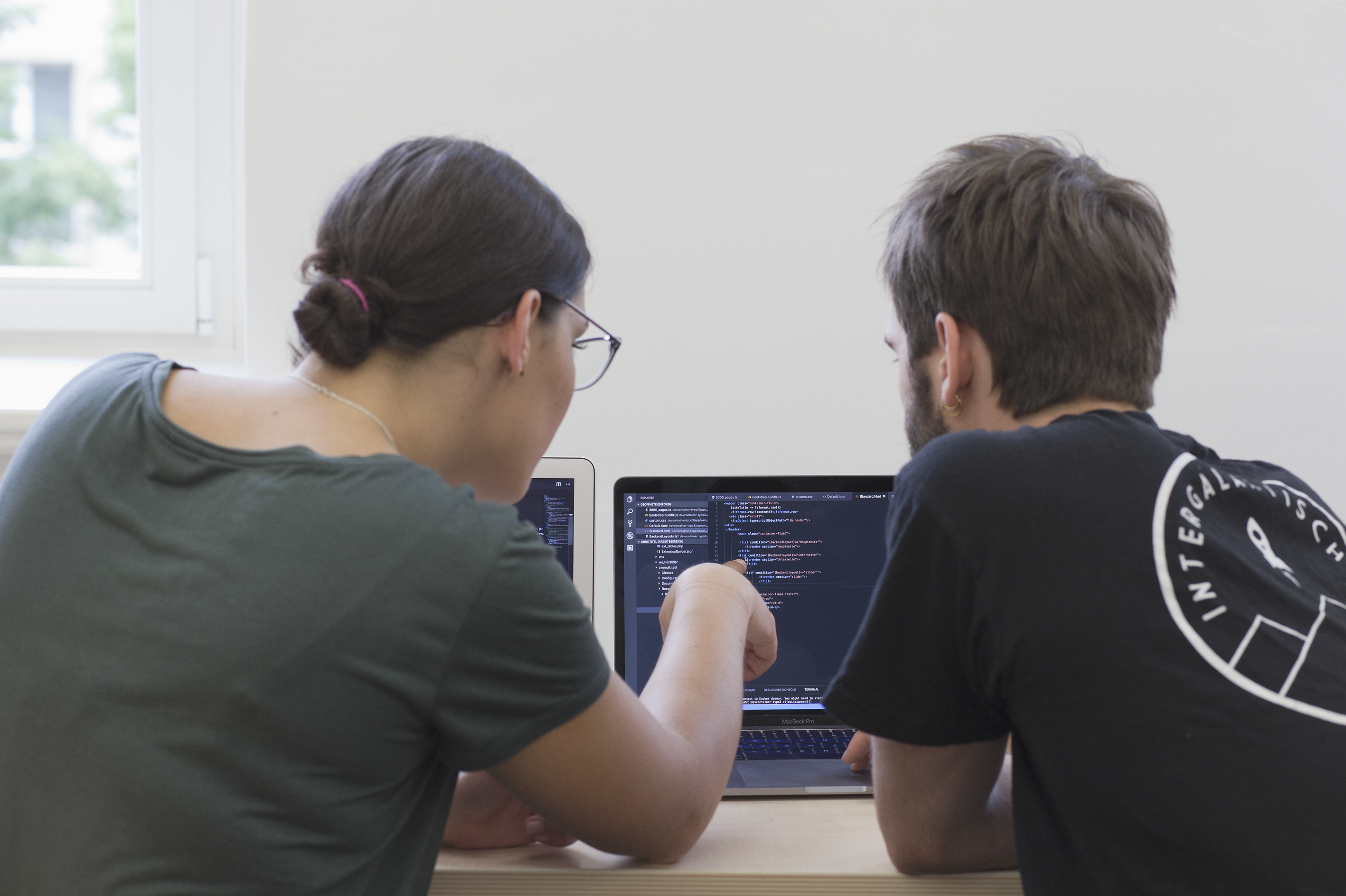 Eine Studentin und ein Student sitzen vor ihren Laptops und schauen sich gemeinsam einen Code auf dem Bildschirm an.