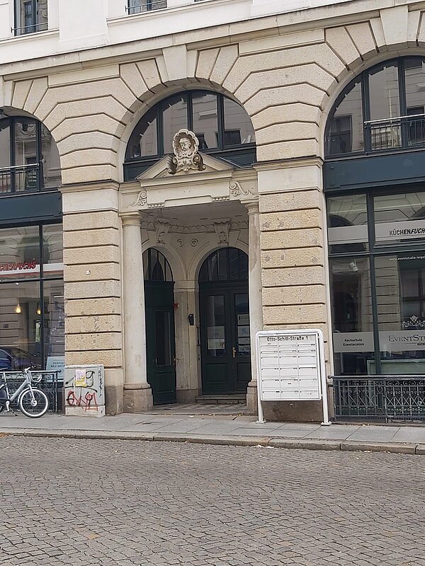 Das Bild zeigt den Eingangsbereich eines Gebäudes, das das Konfuzius-Institut Leipzig beherbergt. Die Architektur wirkt klassisch mit neoklassizistischen Einflüssen. Die Fassade ist in einem sandfarbenen Stein gehalten, und der Eingang wird von großen, eleganten Rundbögen eingerahmt. Über dem Eingang befindet sich ein dekoratives Element, das eine Skulptur oder ein Relief darstellt, welches in das Tympanon eingefasst ist. Dieses Detail verleiht dem Gebäude einen markanten und traditionellen Charakter.