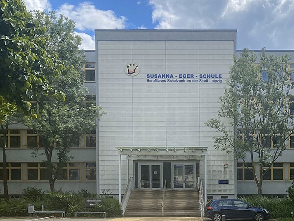 Quaderförmiges Gebäude mit fensterlosem Vorbau, in dem sich der Eingang befindet. Die Fassade ist schmutzig weiß. Eine Treppe führt zum Eingang und der Name des Beruflichen Schulzentrums steht in blauer Schrift auf der Fassade des Vorbaus. Bäume verdecken links und rechts des Vorbaus große Teile des Gebäudes.