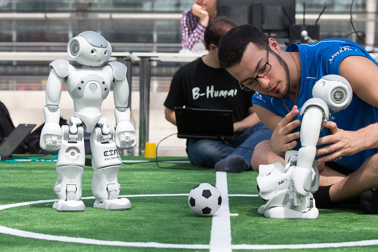 Zwei Fußballroboter beim Anstoß zu einem Fußballspiel der Roboter-Weltmeisterschaft in der Leipziger Messehalle. Ein Mitglied des HTWK-Teams unternimmt letzte Einstellungen an dem 50 Zentimeter großen Roboter.