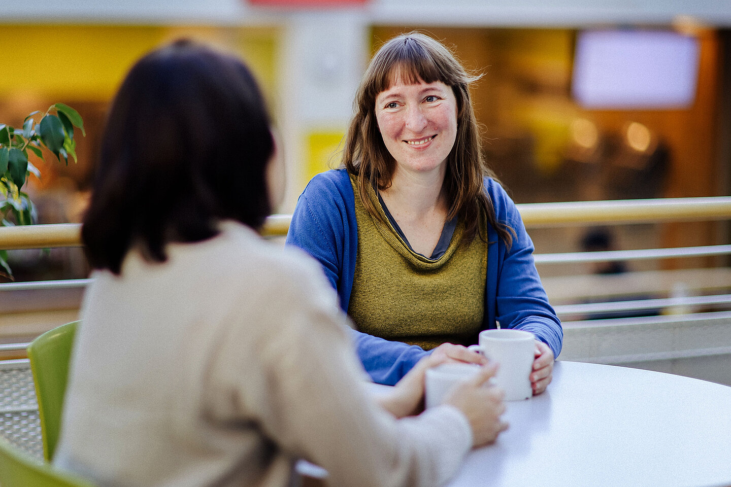 Frau mit Kaffeetasse Eine Frau mit braunen Haaren und blau-grünem Oberteil sitzt an einem Tisch, lächelt und hält eine weiße Tasse in der Hand.