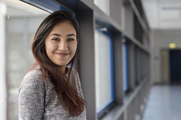 Elaha Fakhri steht in einem Gang der HTWK Leipzig und lehnt an einer Fensterbank. Sie blickt lächelnd in die Kamera.