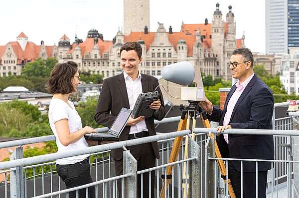 drei Personen messen etwas auf einem Dach, im Hintergrund das Neue Rathaus Leipzig