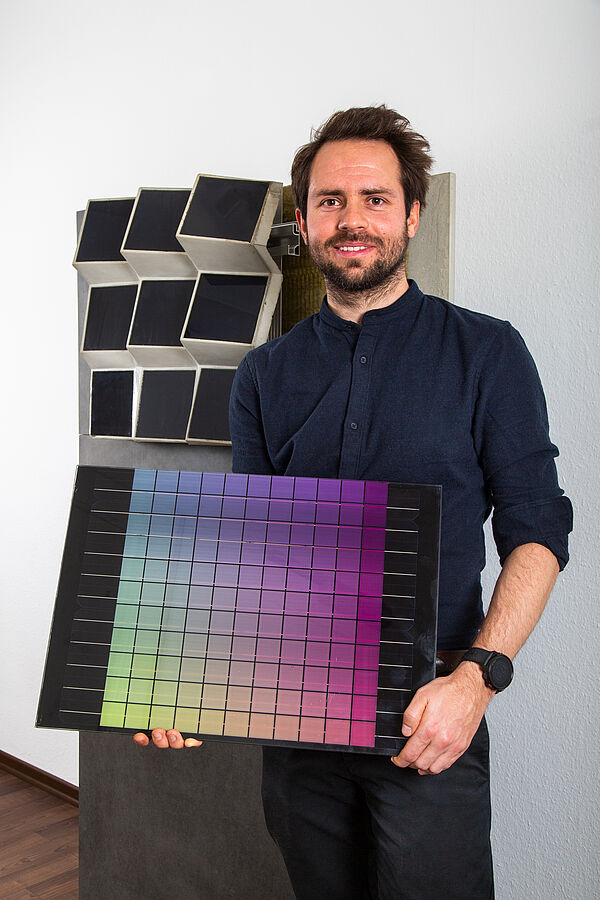 Adrian Heller hält ein Solarmodul in der Hand, bei dem die Farben von gelb bis rosa verlaufen. Er steht vor einem Modell der Solarcon