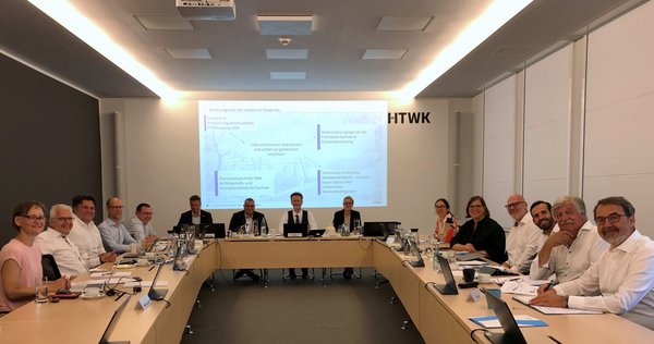 15 Personen sitzen an einem U-förmigen Tisch in Arbeitsatmosphäre