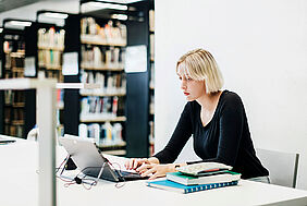 Eine Frau sitzt in der Bibliothek und arbeitet am Laptop.