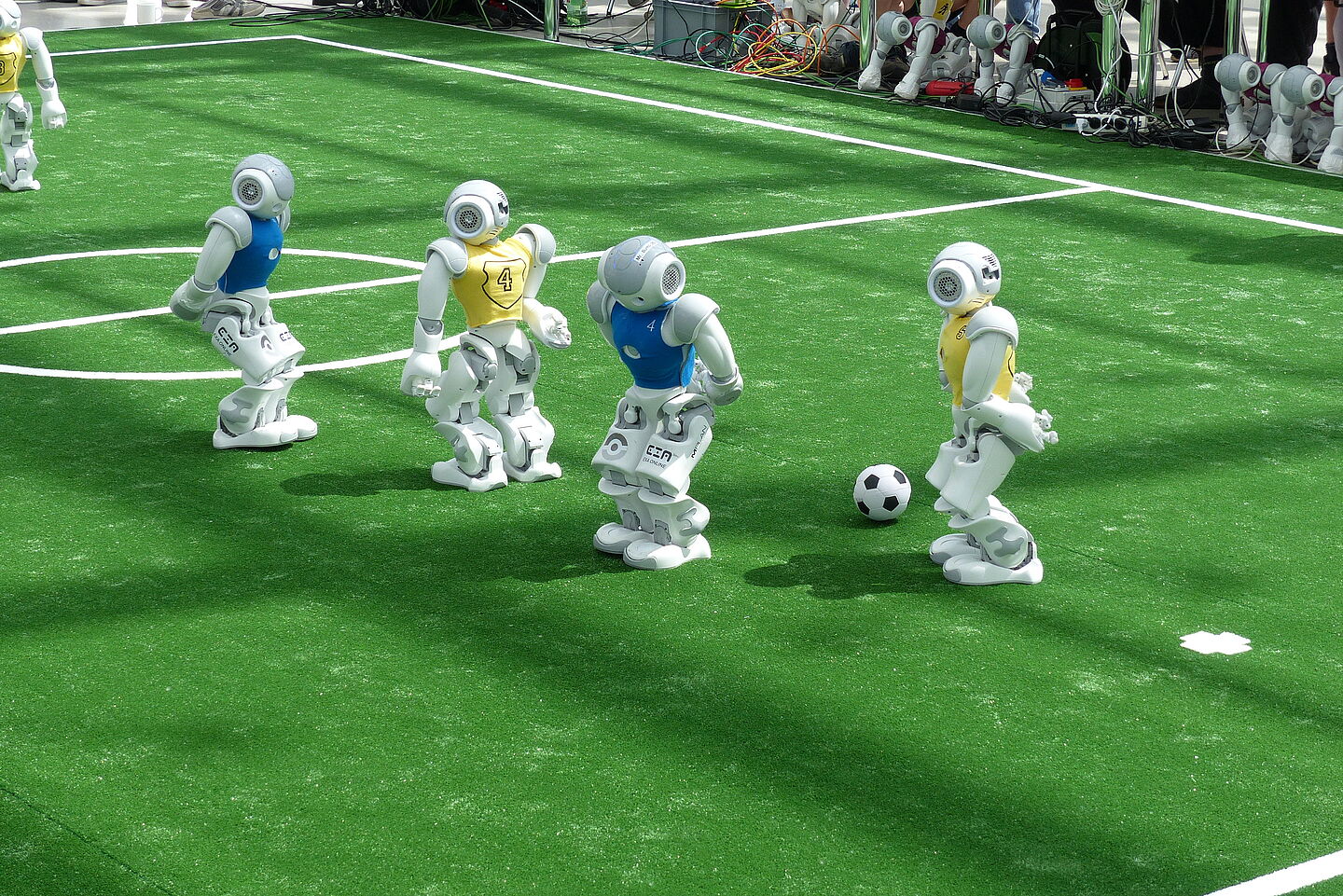 Fußballspiel der Nao-Roboter (Foto: Leipziger Messe/Tom Schulze) Auf einem Fußballfeld stehen vier Roboter in gelben und blauen Trikots. Sie versuchen, den Ball zu spielen.