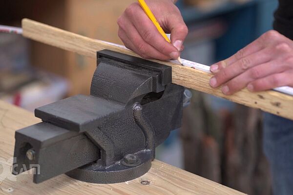 Schraubstock, der ein Stück Holz einfasst. Jemand misst das Stück Holz und markiert mit Bleistift