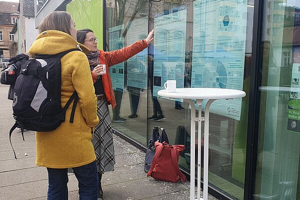 Frau erklärkt anderer Frau Poster, das hinter einer Scheibe hängt.