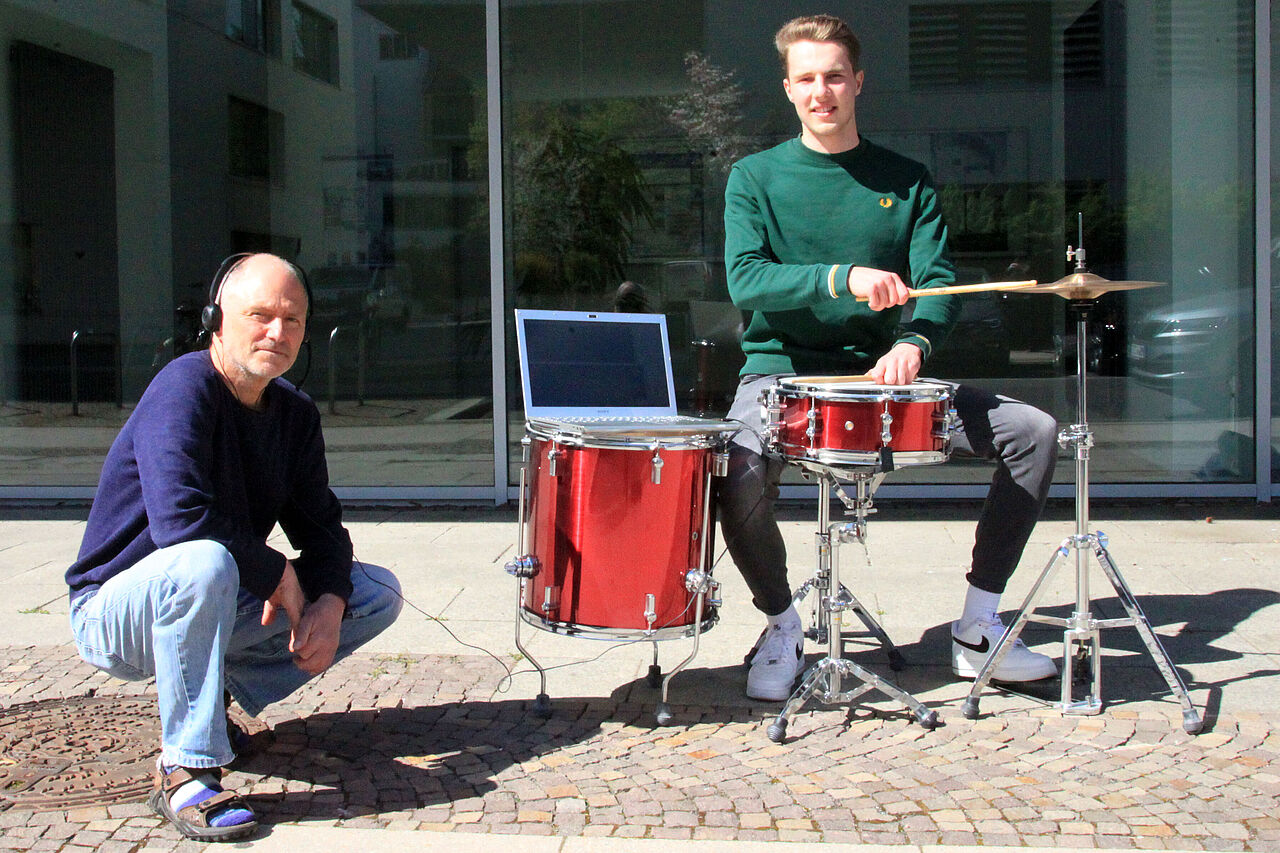 In der Sonne steht ein rotes Schlagzeug, daran sitzt Florian Wedhorn und hält die Schlagzeugstöcke auf die Trommeln; auf einer der Trommeln steht ein Laptop und daneben hockt Professor Christian Weickhardt mit Kopfhörern