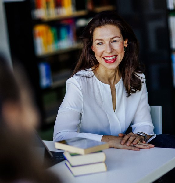 Frau sitzt an einem Tisch mit Büchern in der Hochschulbibliothek und lacht eine gegenübersitzende Person an. 