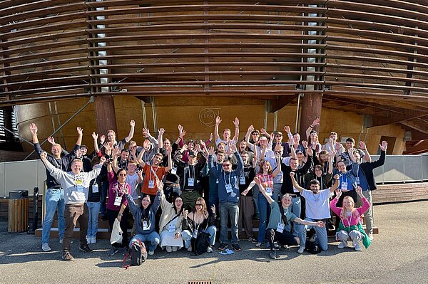 Eine Gruppe freudig winkender junger Menschen vor dem Haupteingang des CERN.