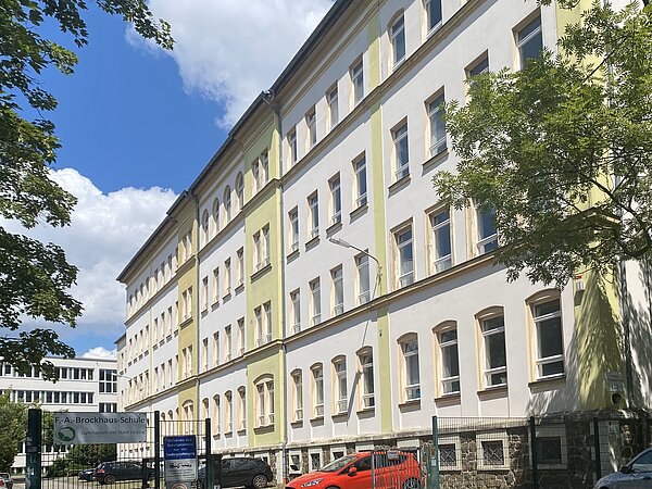 Außenansicht eines vierstöckigen Gebäudes mit weißer Fassade und gelben Details. Davor befindet sich ein Parkplatz mit einigen Autos, der eingezäunt ist.