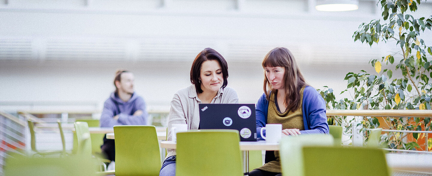 Gemeinsame Laptop-Arbeit Zwei Frauen sitzen nebeneinander an einem Tisch mit einem aufgeklappten Laptop, beide schauen konzentriert auf den Bildschirm.