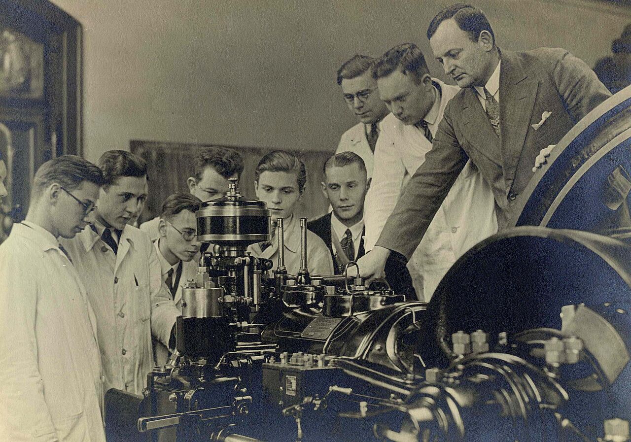 Ältere Fotografie von Studenten, die zusammen mit einem Professor an einer Maschine experimentieren.