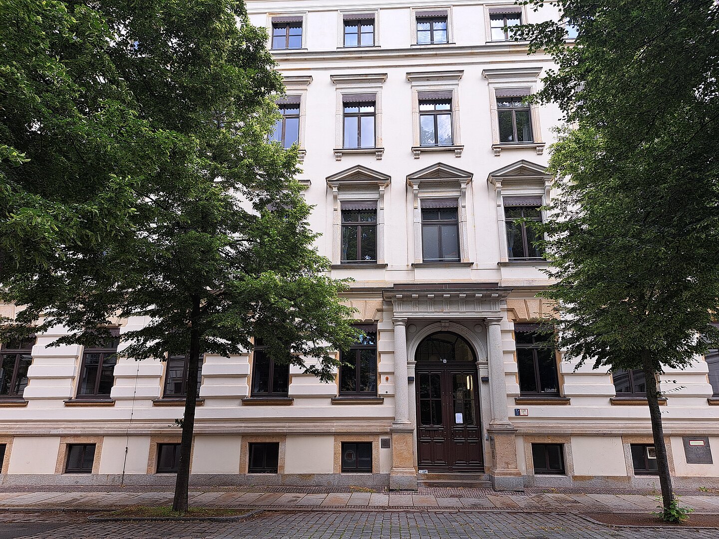 Das Bild zeigt die historische Bibliothek des Thomanerchors in Leipzig, ein elegantes, mehrstöckiges Gebäude mit einer weißen Fassade. Charakteristische Merkmale sind die sorgfältig gestalteten Fenster mit steinernen Einfassungen und Giebeln, die über jedem Fenster angebracht sind. Der Haupteingang ist durch einen Bogen und eine Doppeltür hervorgehoben. Das Gebäude steht in einer ruhigen Straße, umgeben von reichlich Grün durch Bäume.