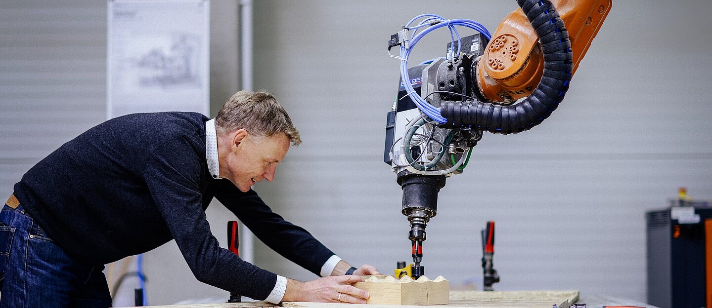 Arbeit an der Industrieroboterarm-Fräse Person beugt sich in einer Werkhalle des HTWK-Holzbauforschungszentrums über eine Werkbank und arbeitet mit einer großen Industrieroboterarm-Fräse