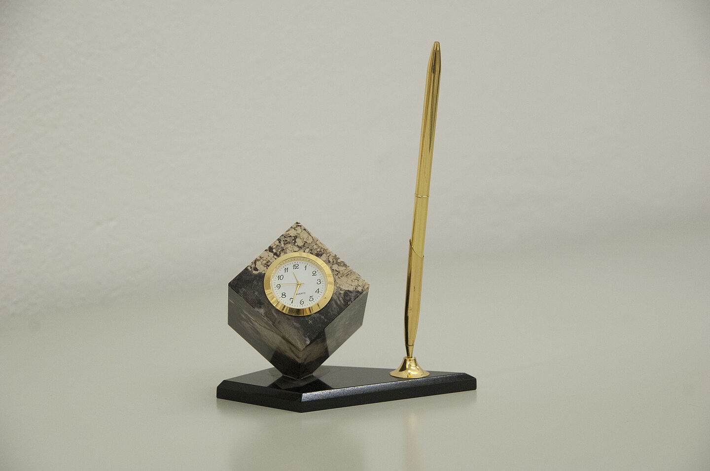 Uhr-Stift-Kombination aus dem HTWK-Archiv (Foto: HTWK Leipzig) Zu sehen ist ein kleiner Würfel aus Marmor, der auf einer seiner Ecken steht und auf einer flachen Steinplatte befestigt ist. In die nach oben zeigende Seite des Würfels ist eine kleine Uhr eingelassen. Neben dem Würfel steht hochkant auf der Platte ein goldener Stift.