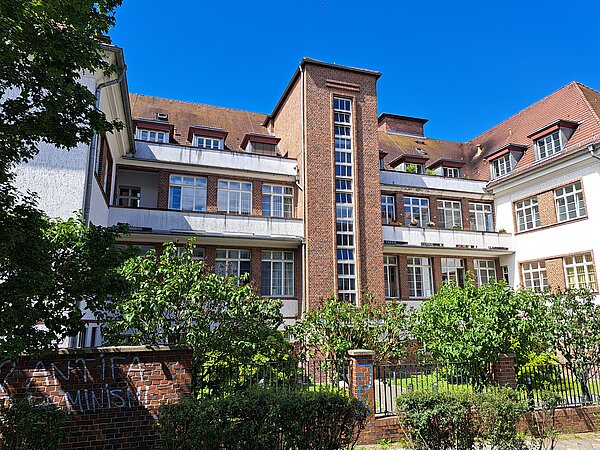 Ein mehrstöckiges Gebäude mit weißer Fassade, die zum Teil aber auch rot verklinkert ist. Das Gebäude hat in jedem Stock kleine Balkone. Vor dem Gebäude ist eine kleine Mauer mit Zaun und einige grüne Büsche.