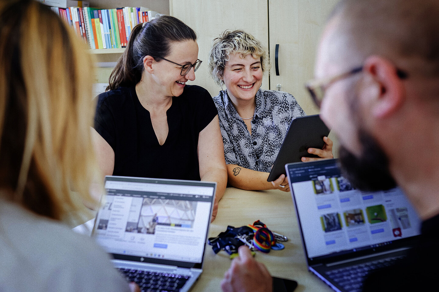 Zwei Frauen sitzen an einem Tisch, eine hält ein Tablet, im Vordergrund sind zwei Personen mit geöffneten Laptops zu sehen.