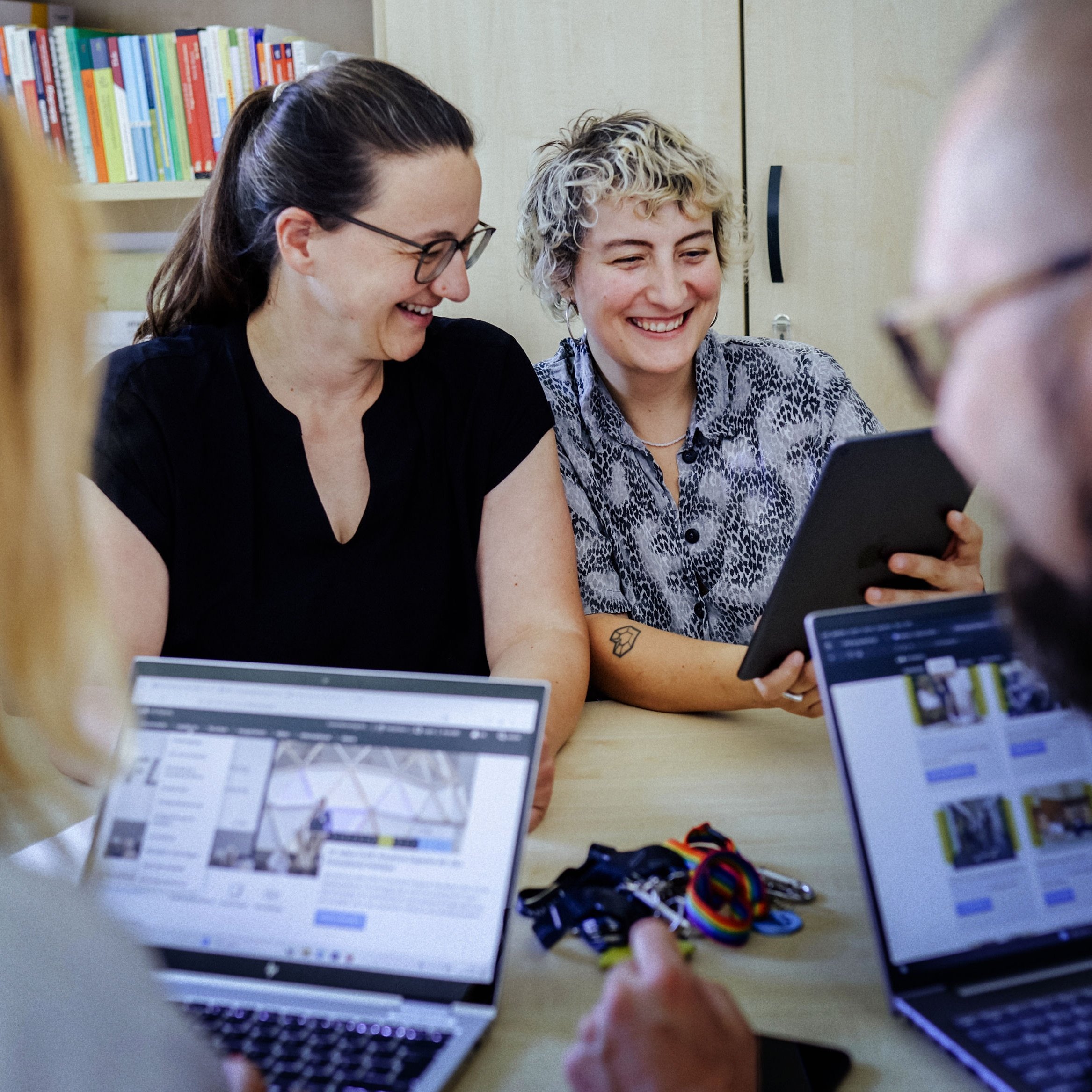 Zwei Frauen sitzen an einem Tisch, eine hält ein Tablet, im Vordergrund sind zwei Personen mit geöffneten Laptops zu sehen.