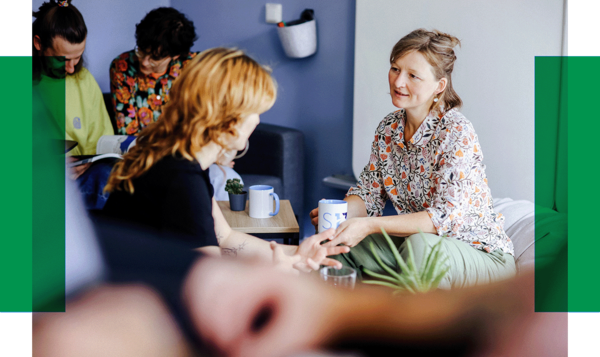 Fünf Personen sitzen in einem hellen Raum, unterhalten sich und trinken Kaffee. Zwei Frauen sprechen sich direkt gegenüber.