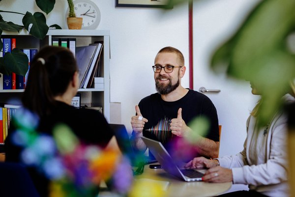 Foto: Anne Schwerin Motivation wächst im gemeinsamen Austausch Ein Mann mit Brille sitzt an einem Tisch und zeigt Daumen hoch, während er mit zwei Personen spricht. Im Hintergrund sind Bücherregale und Pflanzen zu sehen.