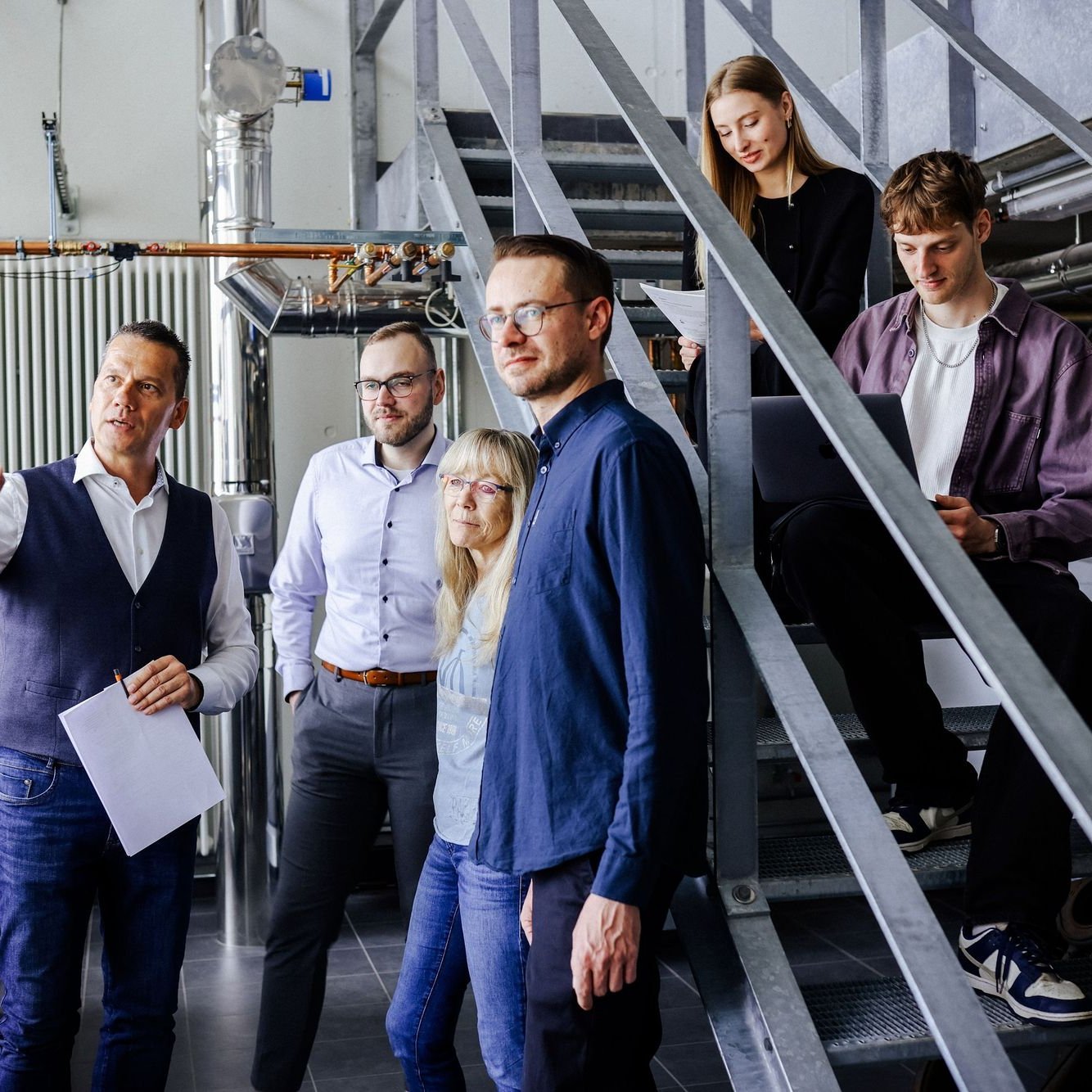 Gruppenbesprechung im Technikbereich Sechs Personen stehen und sitzen neben technischen Anlagen auf einer Metalltreppe, ein Mann vorne erklärt gestikulierend etwas, andere hören oder arbeiten am Laptop.