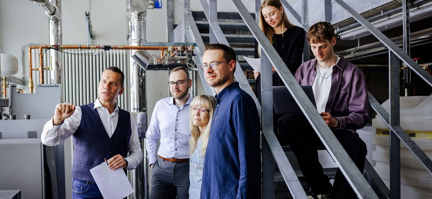 Sechs Personen stehen und sitzen neben technischen Anlagen auf einer Metalltreppe, ein Mann vorne erklärt gestikulierend etwas, andere hören oder arbeiten am Laptop.