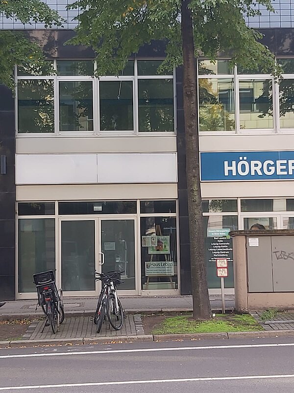 Das Bild zeigt die Fassade eines modernen Gebäudes mit großen Fenstern und einer schlichten Glas-Eingangstür. Links und rechts neben der Tür sind Schilder und Aufkleber angebracht. Auf einem Schild im Eingangsbereich steht „Haus Leben“.