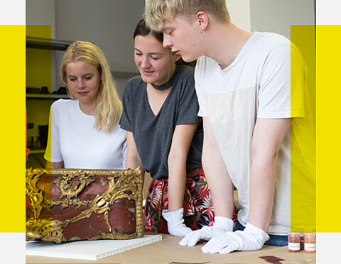 Zwei junge Frauen und ein junger Mann blicken interessiert auf eine vor ihnen liegende antike Kiste.