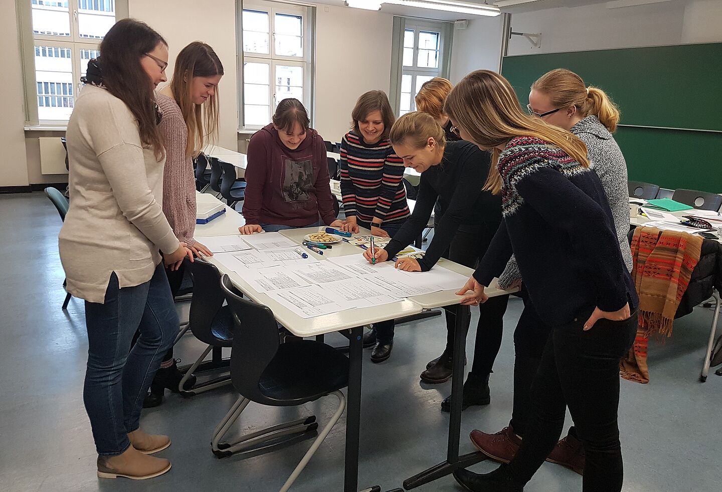 Acht Studentinnen in einem Seminarraum, über einen Tisch mit Papierblättern gebeugt