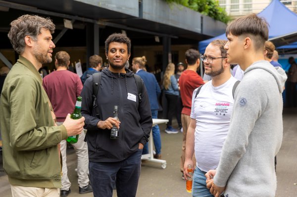 Vier Männer stehen nebeneinander, unterhalten sich und halten jeweils ein Getränk in der Hand. Im Hintergrund sind weitere Gruppen von Menschen zu sehen