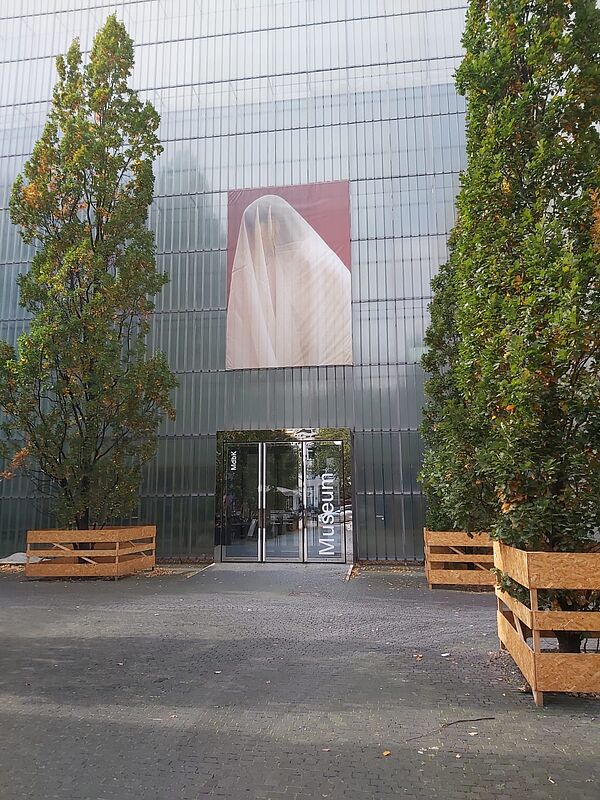 Das Bild zeigt den Eingang eines modernen Museums, das „Museum der bildenden Künste Leipzig“. Die Fassade des Gebäudes ist aus Glas oder durchscheinendem Material, was dem Bauwerk ein minimalistisches und zeitgenössisches Aussehen verleiht. Über dem Eingang hängt ein großes Kunstwerk oder Poster. Der Eingangsbereich ist schlicht gestaltet, mit einer Glasfront, auf der der Schriftzug „Museum“ in weißer Schrift steht. Zu beiden Seiten des Eingangs stehen zwei große Bäume, die von einfachen Holzgestellen eingefasst sind, vermutlich als Schutz. Der Boden vor dem Museum besteht aus grauen Pflastersteinen.