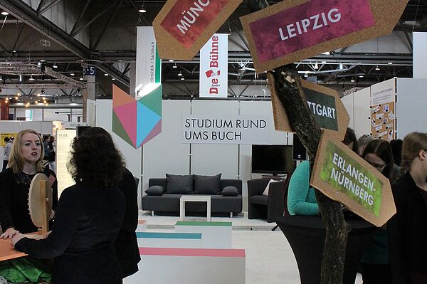 Ein Messestand mit Besucherinnen und Besuchern, vorn ein Wegweiser, im Hintergrund ein Sofa