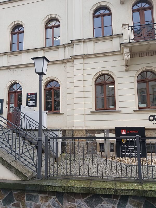 Auf dem Bild ist die Fassade eines Gebäudes zu sehen, das eine klassische Architektur aufweist. Es handelt sich um ein mehrstöckiges Gebäude mit hohen, symmetrischen Fenstern, die bogenförmig sind und braune Rahmen haben. Die Fassade ist in einem hellen Beigeton gestrichen.  Im Vordergrund befindet sich eine Treppe mit einem Geländer aus Metall, das zu einer erhöhten Eingangstür führt.