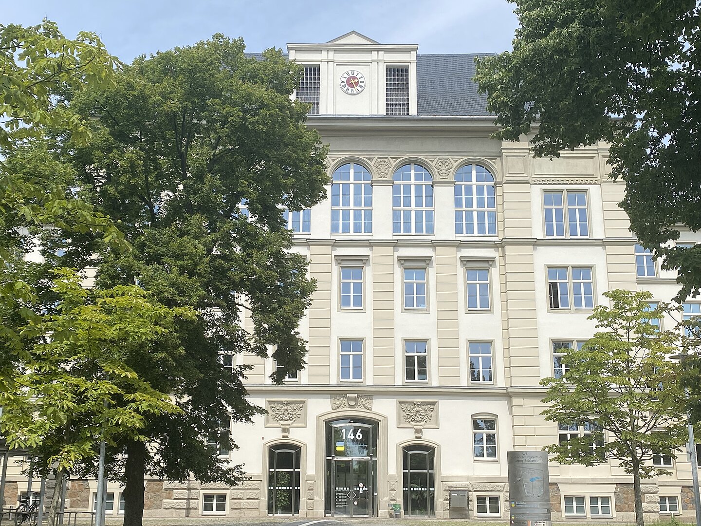 Ein mehrstöckiges Gebäude mit weißen Fassaden und mehrere große Fenster. Der Eingang besteht aus einer großen doppelten Glastür. Auf dem Dach ist eine Uhr zu sehen. Das Gebäude wird von großen Bäumen bedeckt.