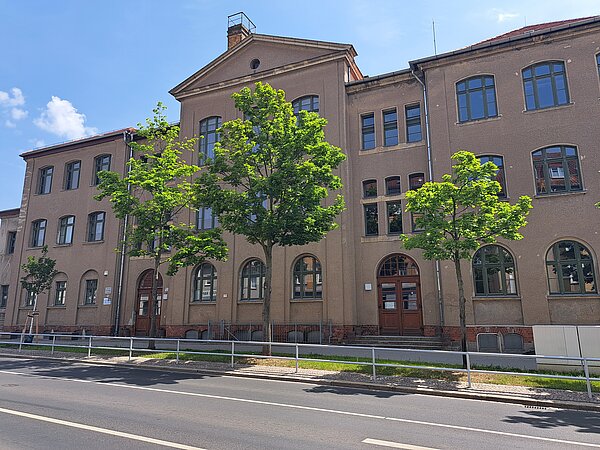 Das Bild zeigt ein braun-graues, mehrstöckiges Gebäude, mit oval abgerundeteten Fenstern und Türen. Es befindet sich an einer Straße und wird teilweise von Bäumen verdeckt.
