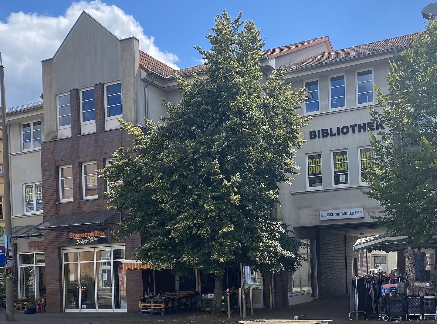 Sie Hochgeladenes Bild beschreibe in 4-5 sätzen Copilot Das Bild zeigt eine Straßenszene mit mehreren Gebäuden und Geschäften. Im Vordergrund steht ein großer Baum, der einen Teil der Gebäude verdeckt. Auf der rechten Seite des Bildes befindet sich ein Gebäude mit der Aufschrift „BIBLIOTHEK“. Darunter ist ein Schild mit der Aufschrift „DANCE COMPANY LEIPZIG“ zu sehen. Links im Bild ist ein Geschäft mit dem Namen „Sternenbäck“ zu erkennen.