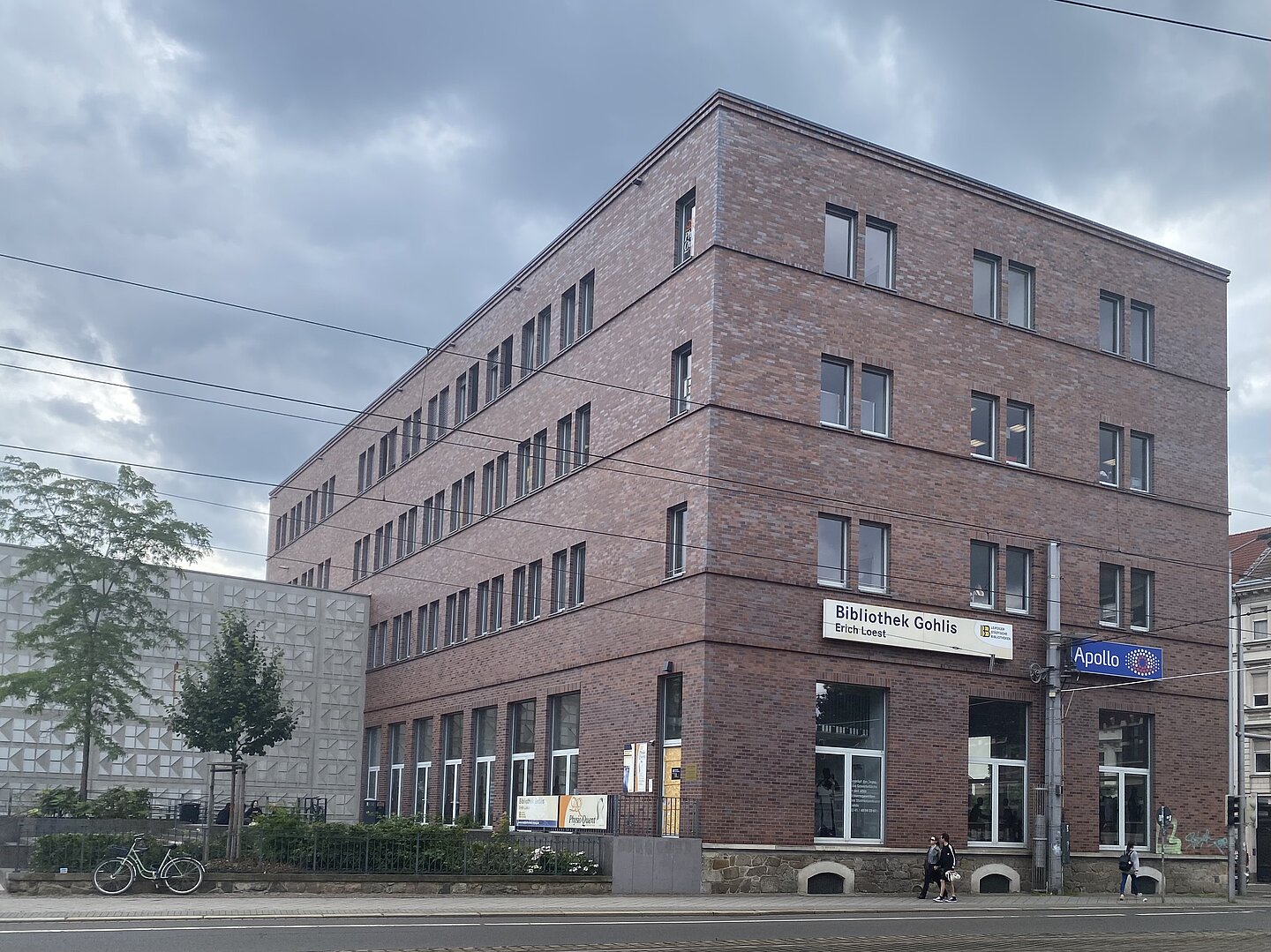 Leipziger Stadtteilbibliothek Gohlis "Erich Loest" Das Bild zeigt ein mehrstöckiges Backsteingebäude mit mehreren Fenstern. Das Gebäude trägt die Schilder „Bibliothek Gohlis Erich Loest“ und „Apollo“. Im Vordergrund sind Straßenbahnlinien zu sehen, und ein Fahrrad ist neben einem kleinen Baum auf der linken Seite des Bildes geparkt.