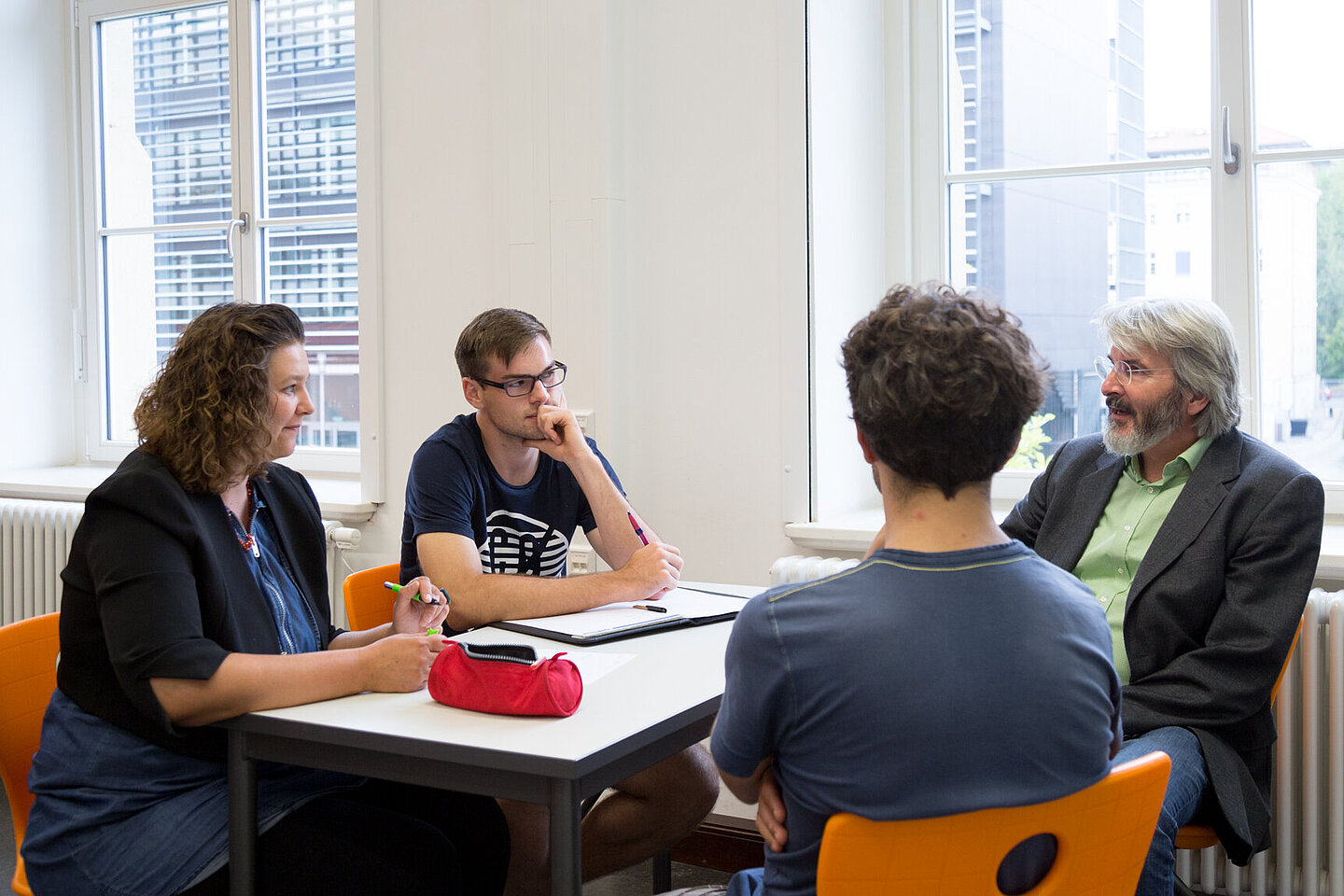 Eine Frau und drei Männer sitzen gemeinsam am Tisch. Ein Mann spricht, während die anderen notieren