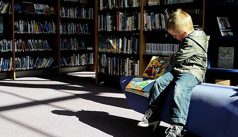 Museumspädagogik Ein Kind sitzt in einer Bibliothek auf einem blauen Sofa und schaut sich ein Buch an.